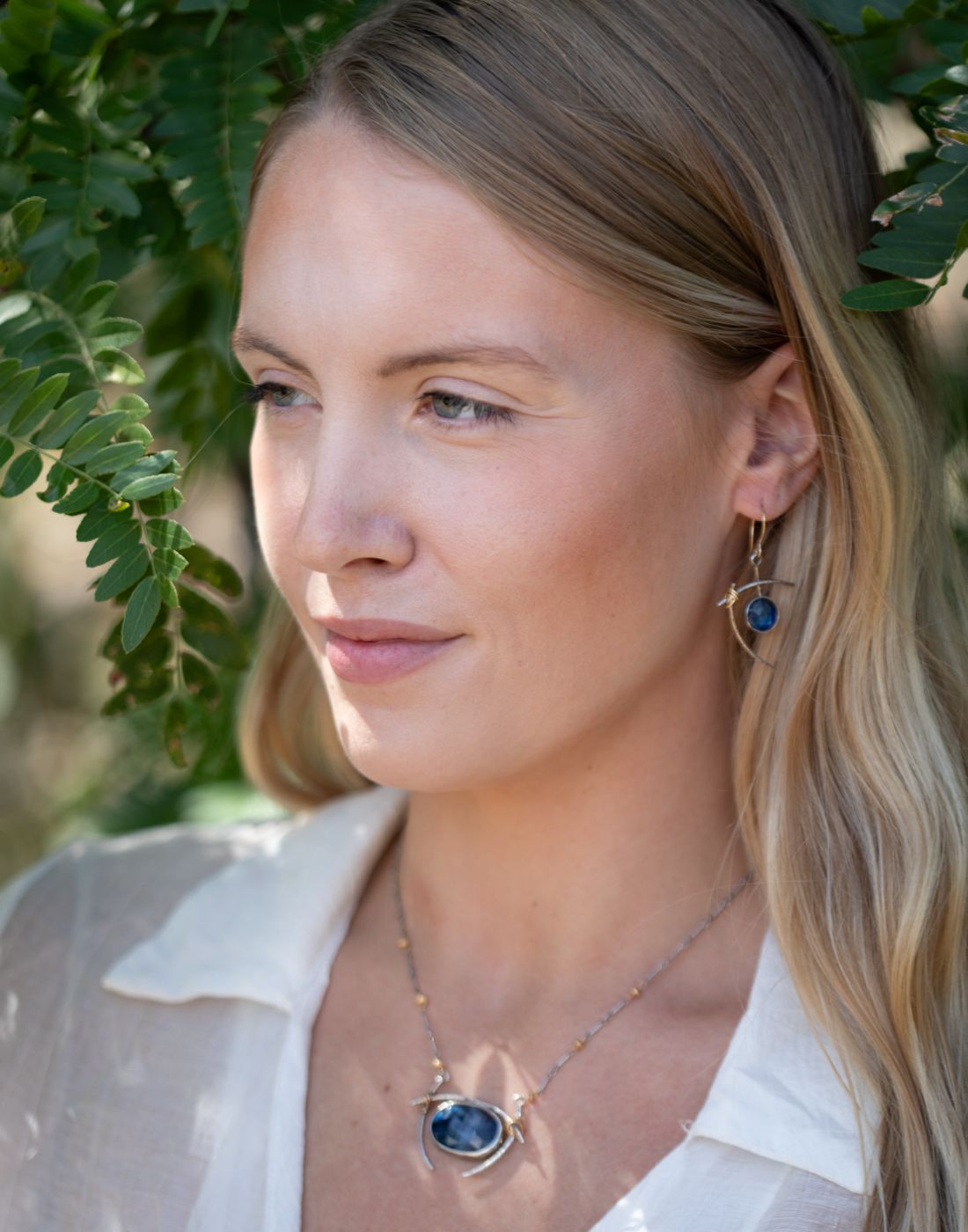 Model wearing nature-inspired handcrafted sterling silver jewelry featuring a necklace and earrings with deep blue gemstones, captured in an outdoor setting surrounded by green leaves.